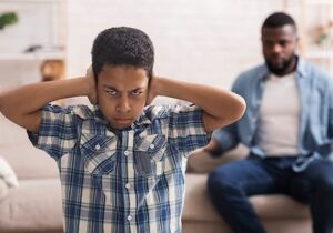 angry teenager covering his ears and facing away from his concerned father is in need of an anger management program